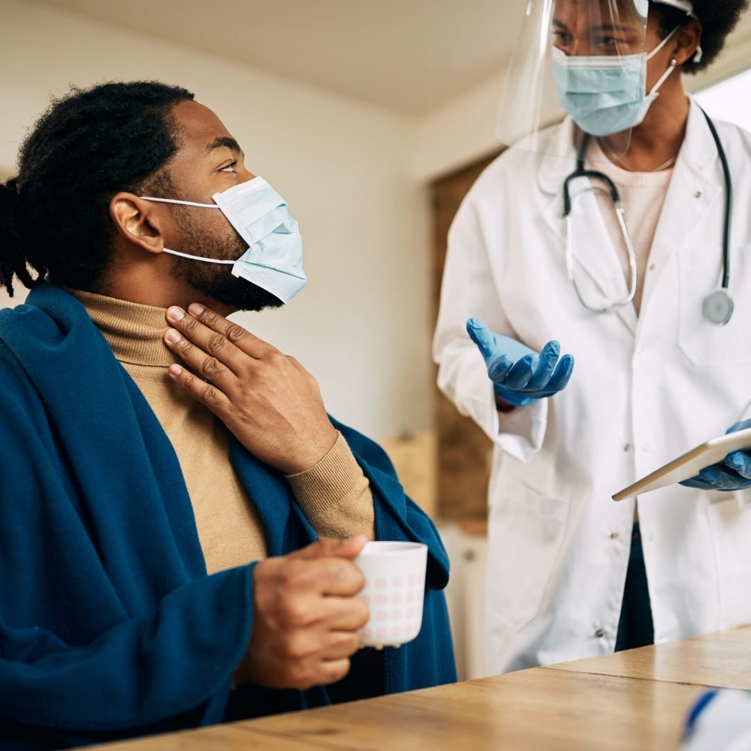 Doctor, I have a very sore throat! African American man wrapped in blanked talking to his doctor and complaining about sore throat during a home visit. Both of them are wearing face masks due to coronavirus pandemic.